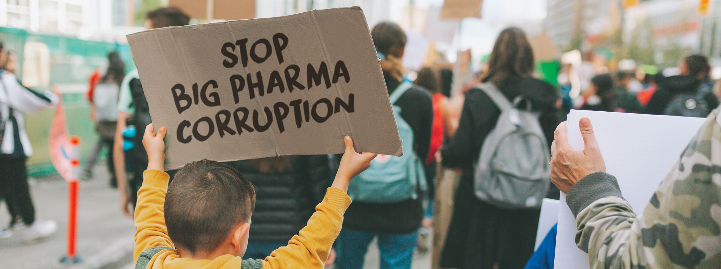 Photo of young boy among other protesters holding a cardboard sign saying Stop Big Pharma Corruption